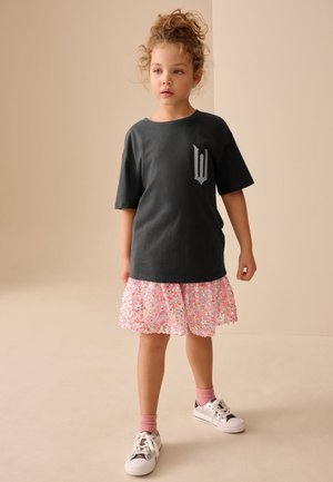 Ung pige med krøllet hår iført en sort oversized T-shirt, pink palietnederdel, pink strømper og hvide sneakers stående mod en beige baggrund.