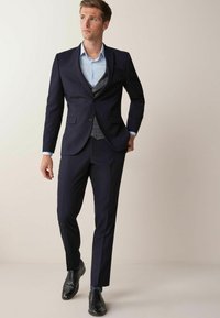 Marinblå kostym med en figursydd kavaj och byxor, ljusblå skjorta och mönstrad väst. Svarta läderskor i formell stil med blå mönstrade strumpor.