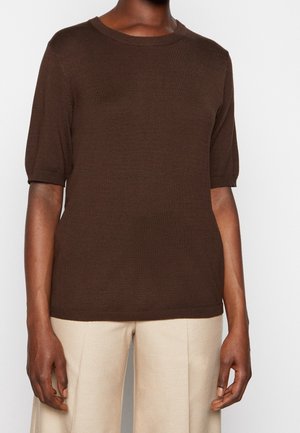 Personne portant un haut en maille manches courtes marron foncé assorti à un pantalon large beige clair sur un fond uni.