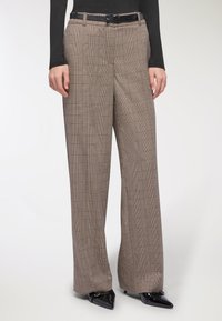 Pantaloni a gamba larga in un motivo a quadri marrone e beige, realizzati in un tessuto texturizzato, con passanti per cintura e indossati con scarpe nere a punta.