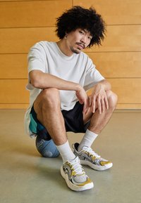 Sneakers met een golvend zwart-wit patroon, geaccentueerd met geel, gedragen met witte enkelsokken. Grijze basketbal op de achtergrond.