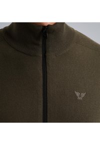Khaki zipsveter s vysokým ribovaným golierom, s čiernym zipsom na prednej strane a malým vyšívaným logom na ľavej strane.