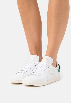 Person trägt weiße Adidas Stan Smith Sneaker mit grünem Fersenlasche und goldener Markenkennung und steht auf einer weißen Oberfläche.
