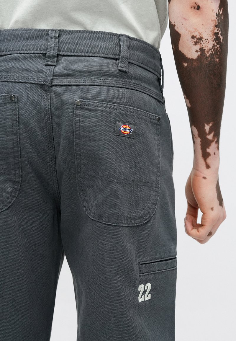 Gray work pants with two back pockets, embroidered number "22," and a logo patch. The fabric appears sturdy with a textured finish.