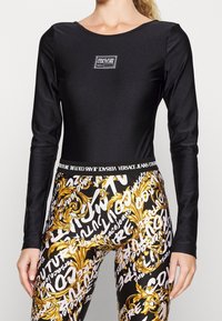 Svart långärmad bodysuit tillsammans med leggings med ett guld- och vittryck, med en distinkt Versace Jeans Couture-logotypdetalj.