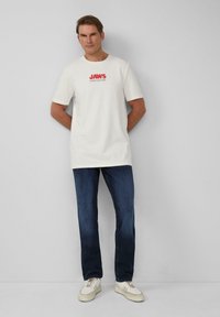 Weißes Baumwoll-T-Shirt mit rotem "JAWS"-Grafikdesign, kombiniert mit dunkelblauen Jeans und weißen Sneakers mit hellbraunen Akzenten.