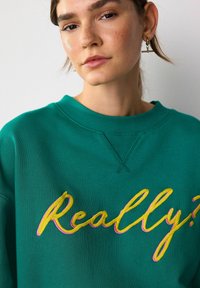 O tânără cu pistrui deschiși și cercei din aur în formă de bară, purtând un hanorac verde brodat cu textul galben și roz "Serios?".