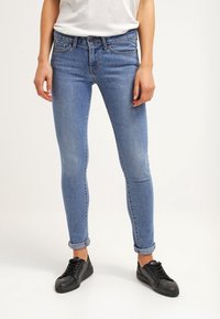 Smala blå jeans av denim med hög midja, klassisk femfickdesign och en blekt finish, stylade med svarta sneakers.