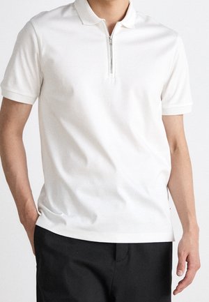 Mand iført en hvid polo med korte ærmer og lynlås foran, kombineret med sorte bukser, stående foran en ensfarvet baggrund.