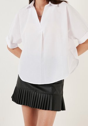 Vit kortärmad skjorta med knappstängning och rak krage, med en längre bakre fåll, som bärs tillsammans med en svart veckad kjol i konstläder.