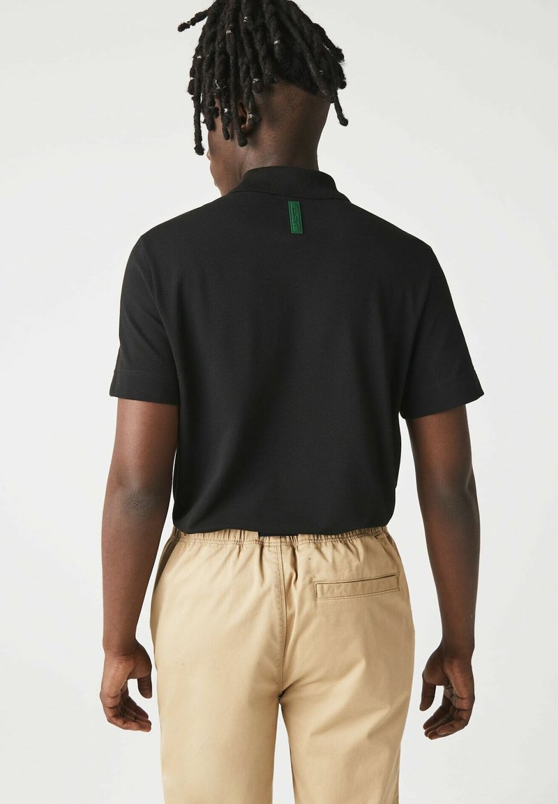 Polo noir avec un col court et des manches courtes, arborant une étiquette verte à l'arrière. Porté avec un pantalon beige à taille élastique.