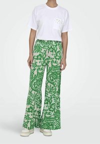 Witte t-shirt met een decoratieve zak, gecombineerd met groene bloemenpatroon wijde broek en witte sneakers.