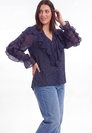 Vrouw met schouderlang bruin haar draagt een marineblauwe blouse met stippen en ruches aan de mouwen en lichtblauwe spijkerbroek, poseert met hand dicht bij het haar.