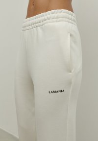 Białe dresy wykonane z miękkiego materiału, z teksturowanym elastycznym pasem w talii i boczną kieszenią, z nadrukiem "LAMANIA" w kolorze czarnym.