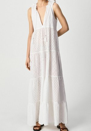 Długa biała sukienka bez rękawów z ażurowym wzorem, głębokim dekoltem w kształcie litery V, marszczonymi ramionami, wielowarstwową spódnicą i frędzlami do wiązania, noszona z brązowymi sandałami.