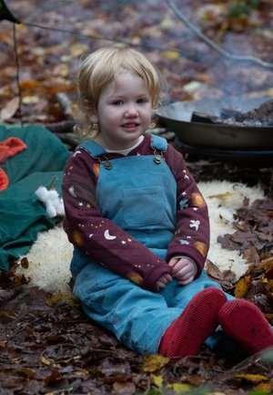 Kind in blauen Corduroy-Latzhosen über einem bordeauxfarbenen, gemusterten Langarmshirt, sitzt auf Blättern, mit roten Socken, umgeben von Natur.