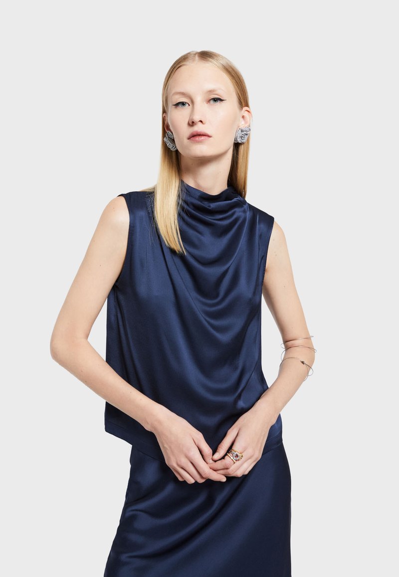 Námornícky modrý saténový top bez rukávov s drapériou okolo krku; hladká textúra, uvoľnený strih, spárovaný so striebornými drôtovými náramkami a prsteňom.