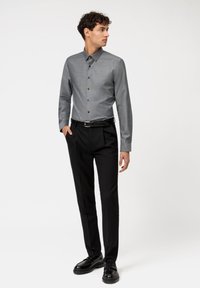 Camicia grigia con bottoni e colletto strutturato, abbinata a pantaloni neri plissettati e scarpe nere, con una texture liscia e una vestibilità sartoriale.