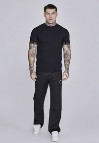 Schwarzes T-Shirt mit kurzen Ärmeln und kontrastierendem goldfarbenem Text auf den Ärmeln, kombiniert mit schwarzen Jogginghosen mit dezentem Logo, getragen mit weißen Turnschuhen.