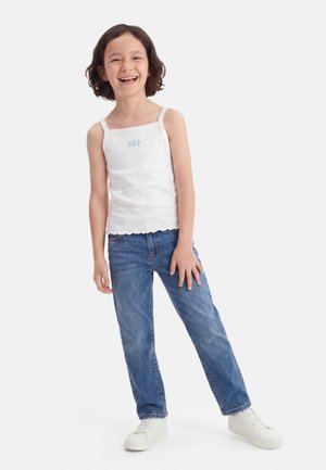 Παιδί που χαμογελά και στέκεται με το ένα χέρι στη λεκάνη, φοράει λευκό αμάνικο μπλουζάκι, μπλε τζιν και λευκά αθλητικά παπούτσια σε απλό φόντο.