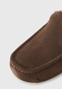Bruine suède slof met een zachte textuur, ronde vorm en zichtbare stiksels langs de rand. Naadloos ontwerp, geen beslag aanwezig.
