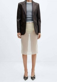 Frau trägt ein schwarzes Lederblazer, einen grauen, kurzen Pullover, einen durchsichtigen beigen Rock mit schwarzen Punkten und schwarze spitze Absätze, steht aufrecht.