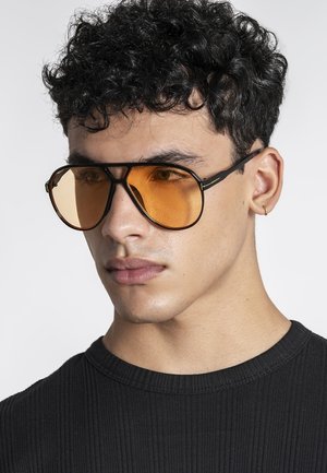 Mladý muž s kučeravými vlasmi, ktorý má na sebe prekročené pilotné slnečné okuliare s oranžovým odtieňom a malé kruhové náušnice, oblečený v čiernom rebrovanom tričku.