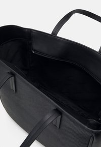 Sac cabas noir texturé avec deux poignées, montrant un intérieur spacieux doublé de tissu de marque et une poche zippée sur le côté.