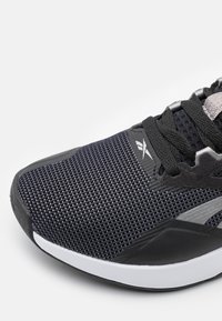Scarpa sportiva nera con upper in mesh testurizzato, accenti metallici e dettagli grigi. Presenta una suola bianca imbottita e lacci neri.