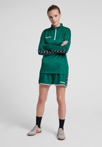 Top sportivo verde con zip e accenti a zigzag neri, abbinato a pantaloni corti verdi con bordi bianchi, indossato con scarpe sportive grigie e calzini neri.