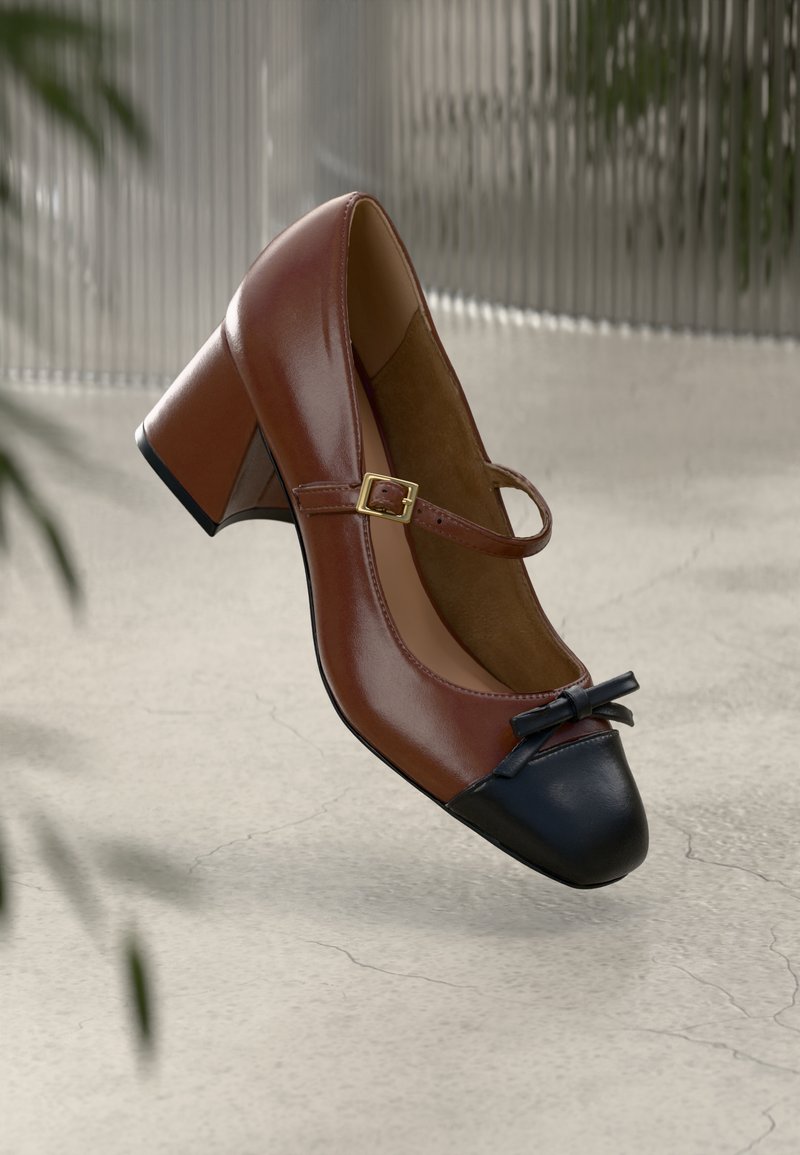 Talons Mary Jane en cuir marron avec bout noir, nœud noir, bride à boucle dorée et talon bloc sur un sol clair texturé.