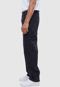 Jeans in denim nero con una vestibilità rilassata, tasche laterali e una texture leggermente sbiadita. Abbinati a sneakers bianche. Vista laterale.