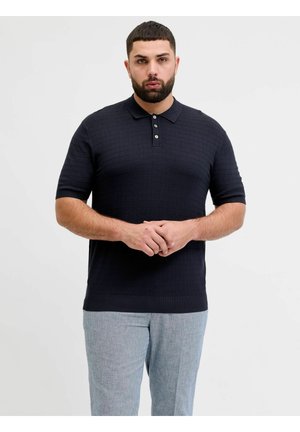 Mand med skæg iført mørkeblå tekstureret polo med korte ærmer og lysegrå bukser, stående med hænderne foldet foran en hvid baggrund.