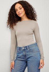 Langärmliges, eng anliegendes Oberteil aus hellbeigem Stoff, kombiniert mit hoch geschnittenen blauen Denimjeans. Glatte Textur mit einheitlicher Farbe.