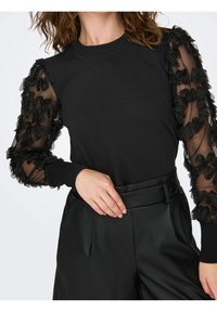 Sudadera negra con mangas de malla negra transparente con volantes, con escote redondo y puños de canalé. Combinada con pantalones negros de talle alto.