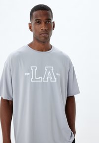 Camiseta gris de manga corta hecha de tela suave, con un gráfico blanco de "LA" y el texto "EST: 1992" en el frente. Corte relajado.