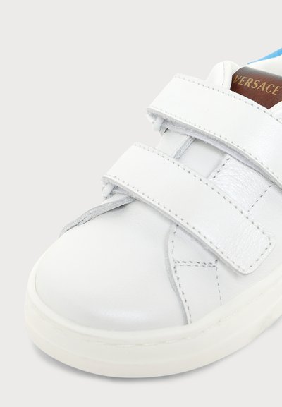 Baskets en cuir blanc avec deux bandes velcro et une étiquette marron montrant une partie du logo Versace sur la languette.