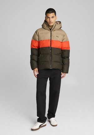 Chaqueta tipo plumón en beige, naranja y verde oscuro. Presenta capucha, cierre de cremallera y puños elásticos. Combinada con pantalones negros y zapatillas blancas.