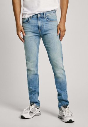 Des jeans en denim bleu clair avec une coupe ajustée, un design classique à cinq poches, un délavage subtil et des coutures contrastées. Portés avec des baskets grises et blanches.