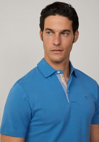 Blå pikétröja i bomull, har en randig knäppning i gul och vit, korta ärmar och en broderad logotyp på bröstet.