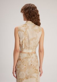 Blusa sin mangas con cuello tipo camisa. Tejido en color beige claro con patrones florales en crema y dorado. Diseño corto con una silueta ajustada.