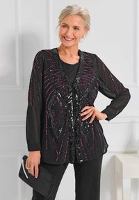 Schwarze transparente Strickjacke mit geometrischem Paillettenmuster in lila und schwarz. Lange Ärmel, V-Ausschnitt, kombiniert mit einem schwarzen Oberteil und einer schwarzen Clutch.