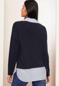 Top din material bleu marinar, cu mânecă lungă și croială relaxată, purtat peste o cămașă deschisă la culoare, cu dungi, care are un tiv rotund și un material texturat.