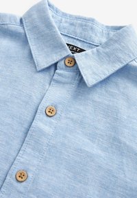 Chemise bleu clair à col avec boutons en bois et une étiquette de marque noire partiellement visible à l'intérieur du col.