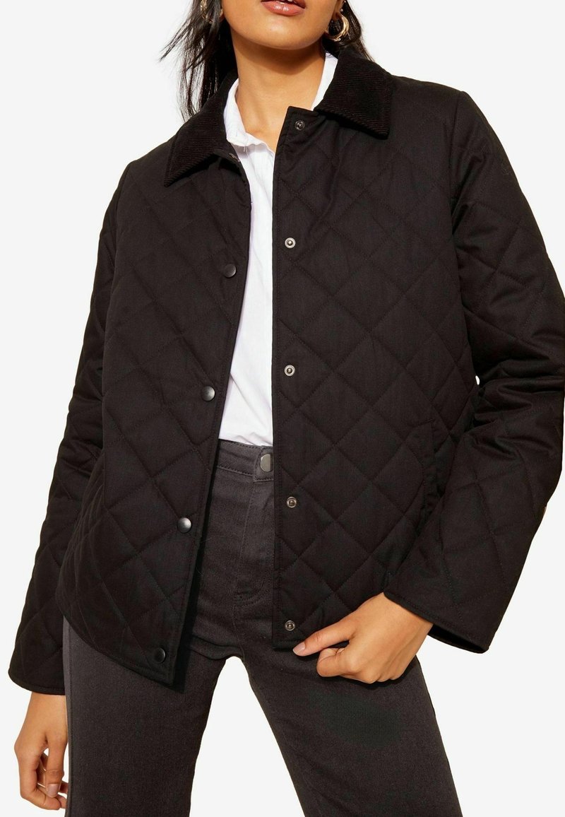 Černá prošívaná bunda se kordovým límcem, cvokovými knoflíky a strukturovanou tkaninou. Nositá přes bílé tričko a tmavé džíny.