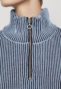 Mock-Neck-Pullover in Blau und Marineblau mit gestreiftem Design, ausgestattet mit einem silbernen Reißverschluss und strukturiertem Strickstoff. Nahaufnahme des Kragens und Reißverschlussdetails.