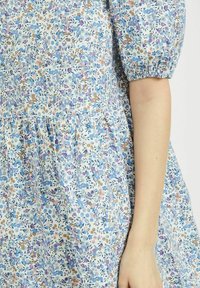 Robe florale bleue avec manches bouffantes, présentant un fond clair et des fleurs multicolores dans des tons de bleu, rose et marron. Texture de tissu douce.