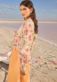 Blusa floreale di un delicato rosa con maniche arricciate, decorata con fiori rossi e gialli. Abbinata a pantaloni arancioni a vita alta e gamba dritta.