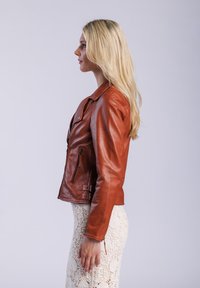 Bruine leren motorjack met ritsen en gespen, met een slim fit silhouet. Model draagt een gestreepte crèmekleurige rok eronder.