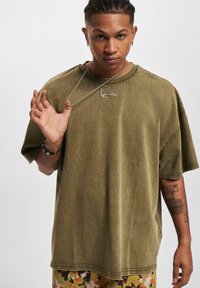 Olive green oversized t-shirt with short sleeves and a ribbed crew neck. Features a silver chain necklace and visible tattoo on arm.